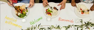 Symbolbild: Auf einem Tisch steht in mehreren Sprachen "herzlich willkommen" darüber sind gefüllte Teller, von den Menschen sieht man nur die Hände